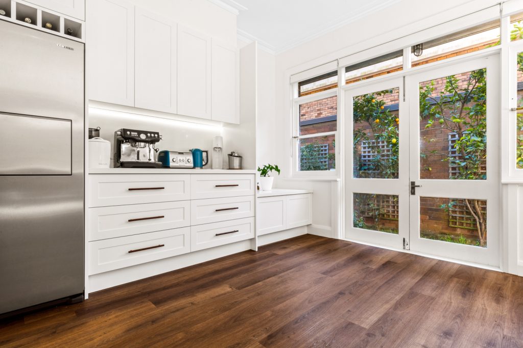 White kitchen cabinetry beside a stainless steel fridge and glazed French doors overlooking the garden.