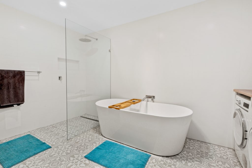 Freestanding bath beside a glass shower screen, with patterned floor tiles and laundry cabinetry at the edge of the room.