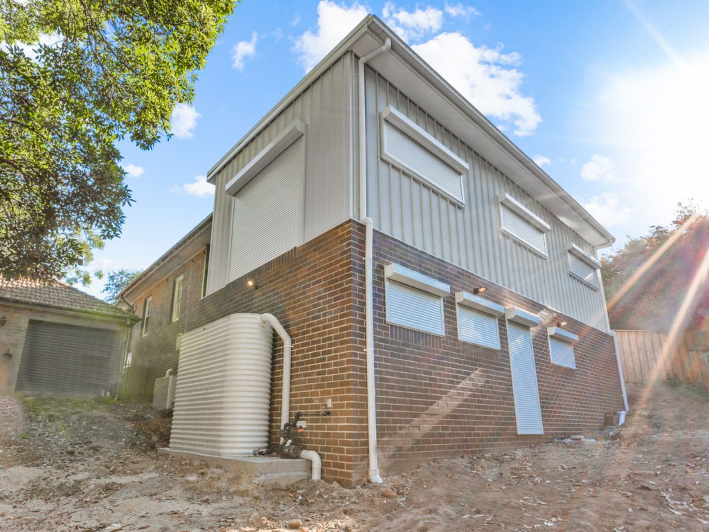 Two-storey rear extension with brick lower walls, vertical upper cladding, closed external shutters and a rainwater tank on the sloping site.